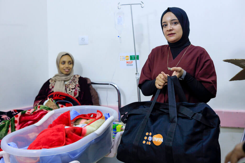 Mother in Gaza poses next to her baby, while her mother in law holds a UNFPA mama kit